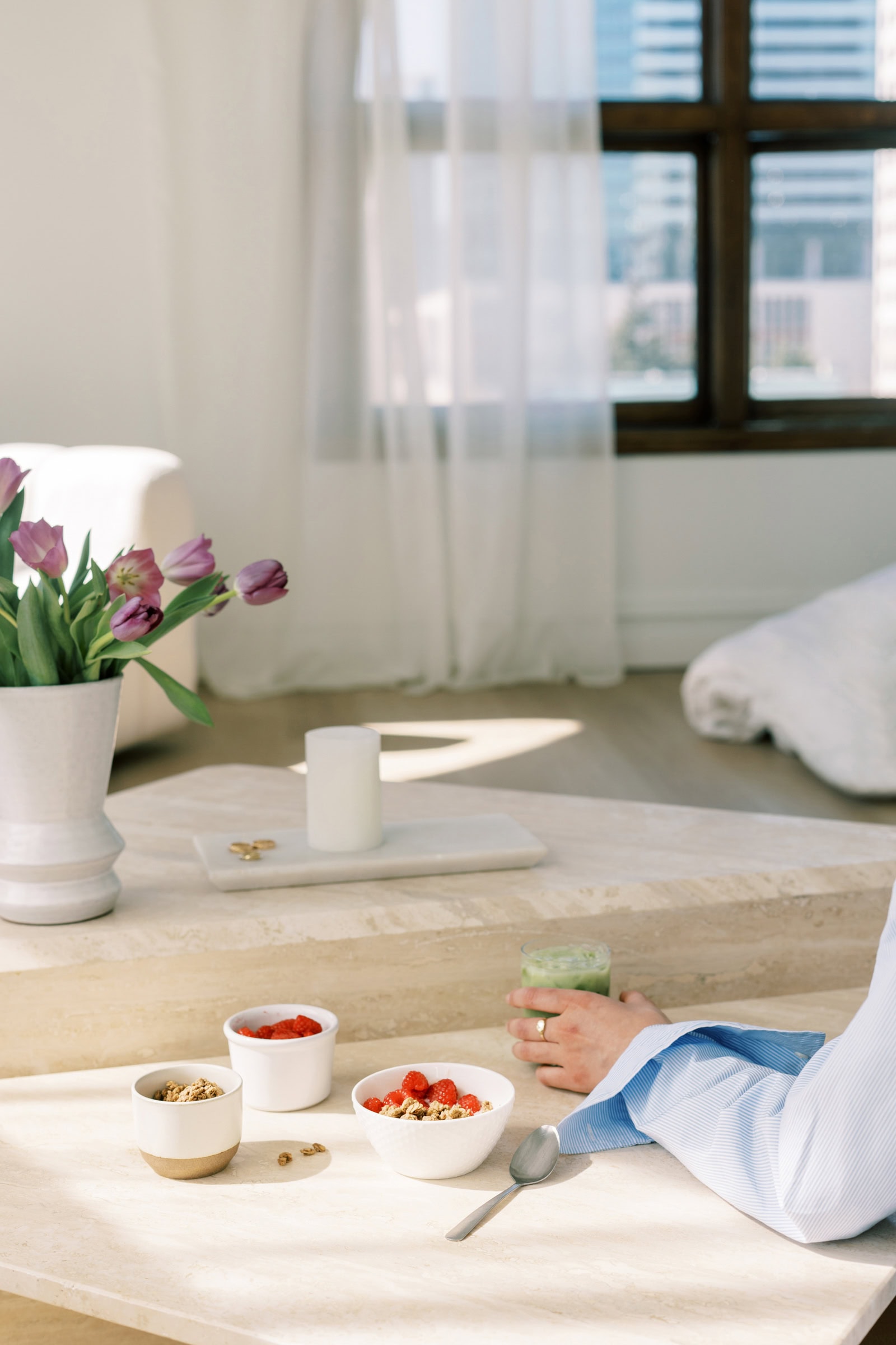 A serene breakfast setup featuring bowls of granola, fresh berries, and a green smoothie on a minimalist marble table. A vase of purple tulips adds elegance to the sunlit space.