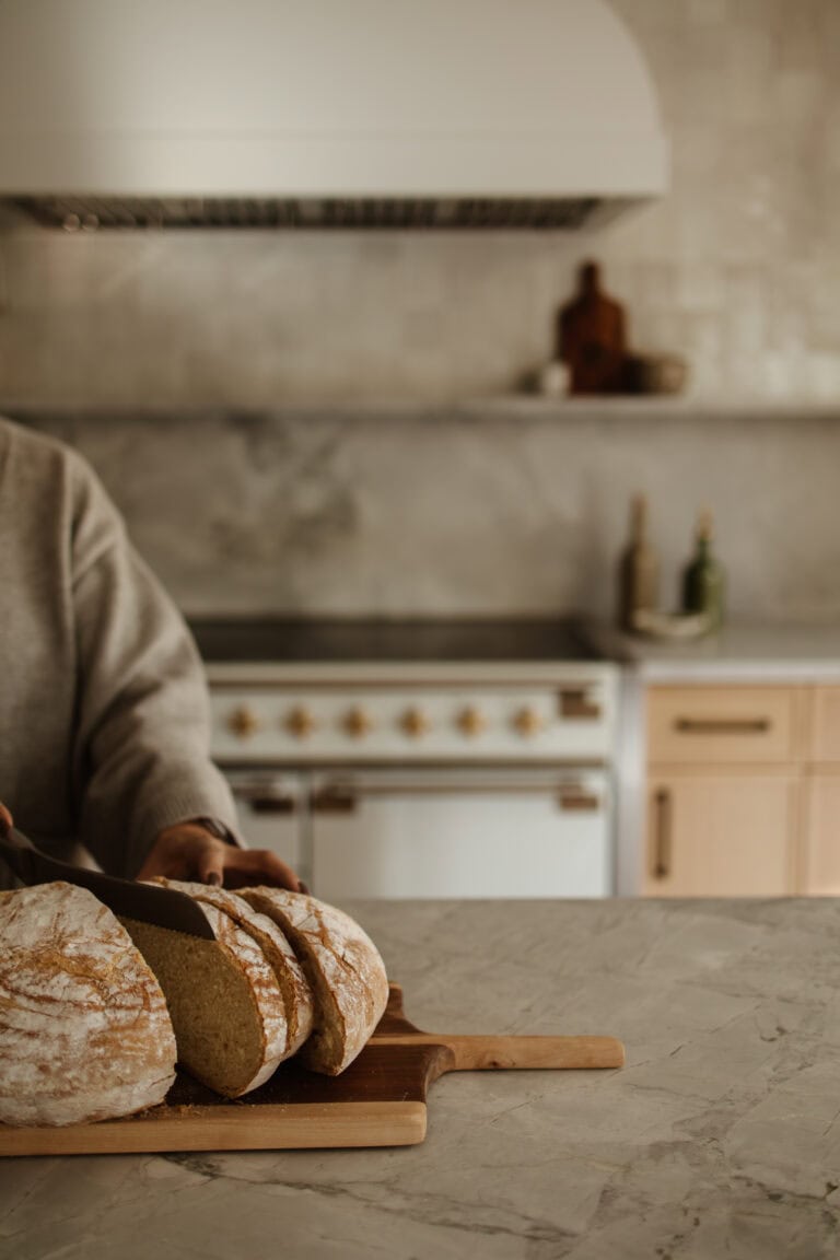 A rustic kitchen with a person slicing a fresh loaf of artisan bread on a wooden cutting board. The backdrop includes a marble countertop and a classic range oven.