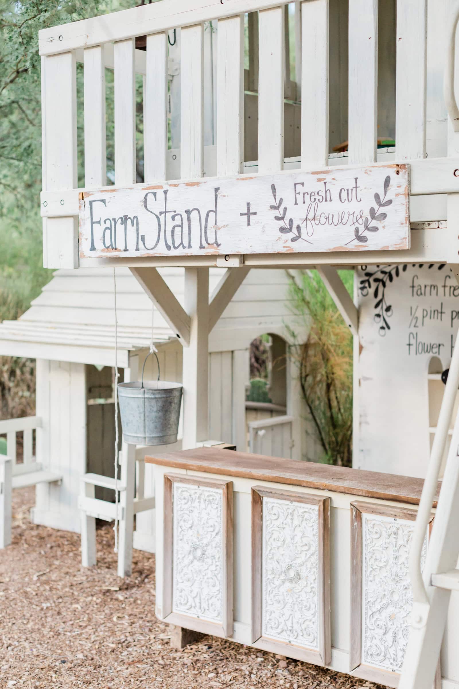 A charming rustic farm stand with white wood panels, ornate detailing, and a sign that reads "Farm Stand + Fresh Cut Flowers" in a whimsical font, surrounded by greenery.