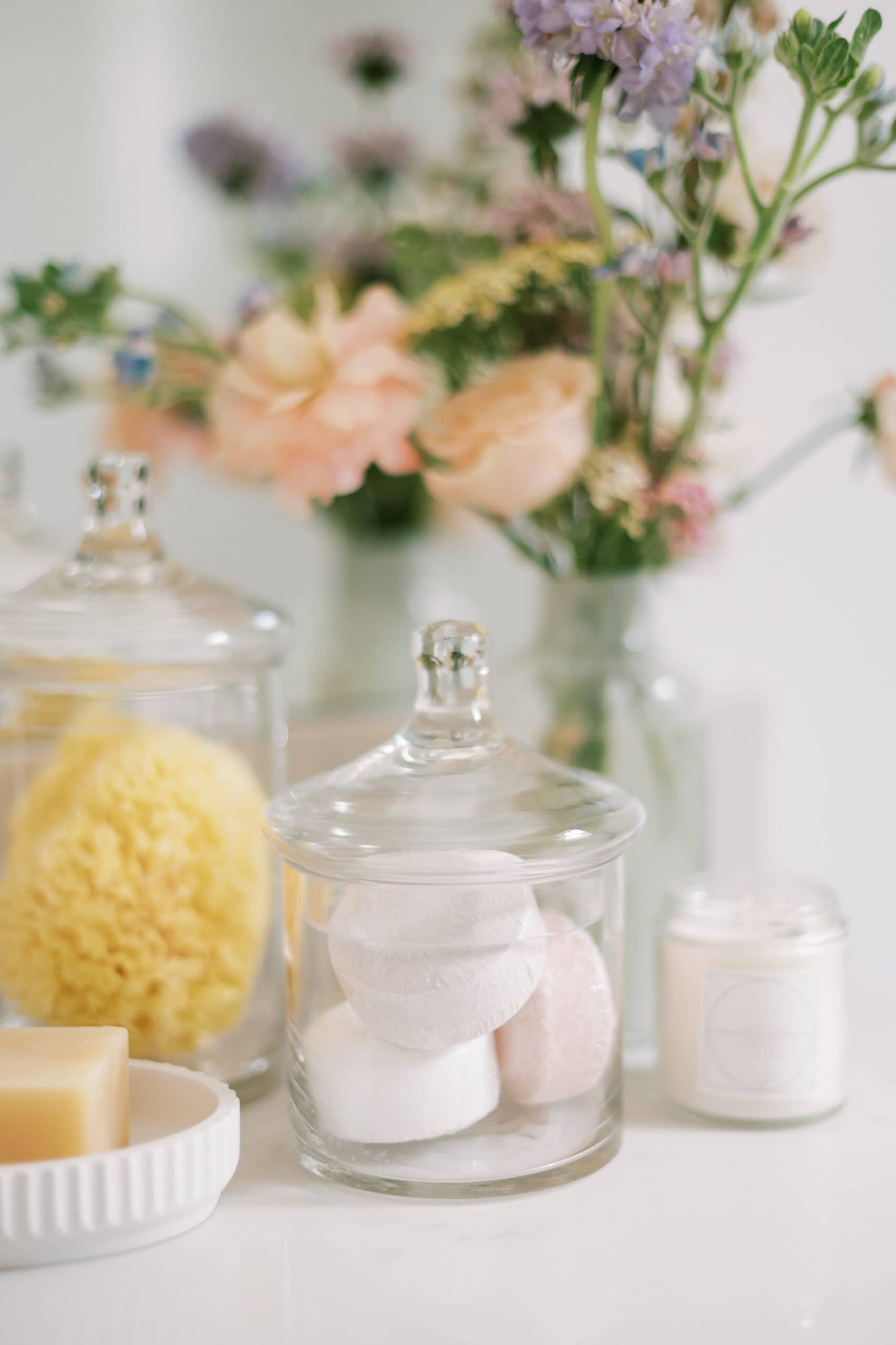 A serene bathroom scene featuring glass jars with bath essentials like sponges and bath bombs, a soy candle, and pastel-colored flowers in the background. The soft focus on the flowers creates a calming and spa-like ambiance.
