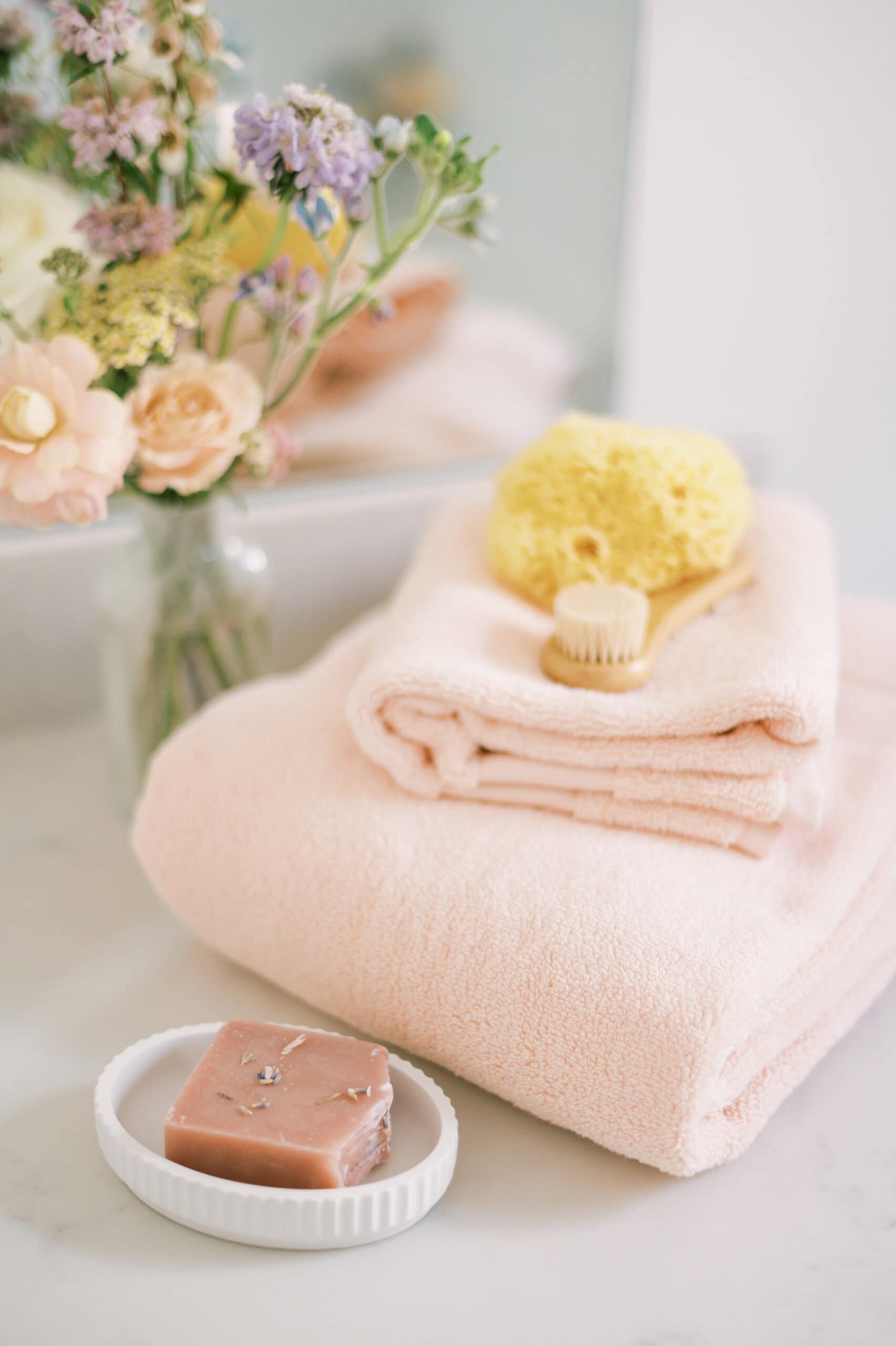 A stack of peach-colored towels on a bathroom counter, accompanied by a natural sea sponge, a wooden body brush, and a bar of soap on a small dish. Delicate flowers in a vase add a fresh and relaxing touch.