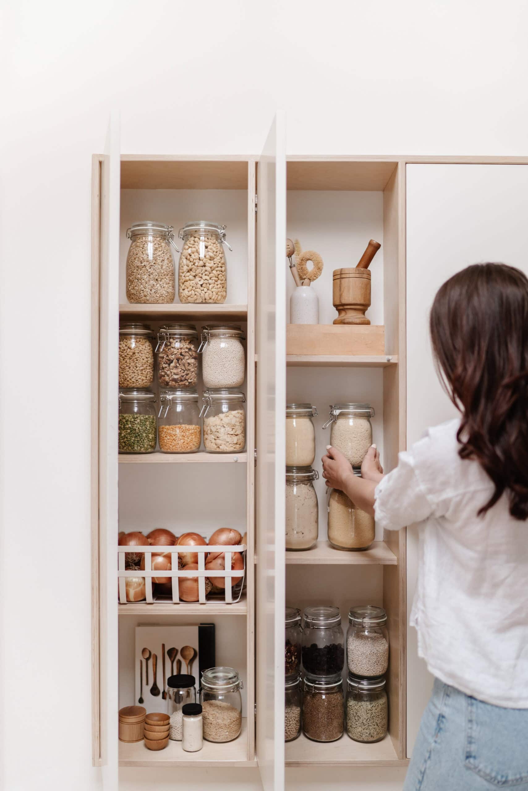 A beautifully organized pantry with jars of grains, legumes, and nuts, along with wooden kitchen tools and a basket of onions. A woman in a white shirt organizes a glass jar on the shelves.