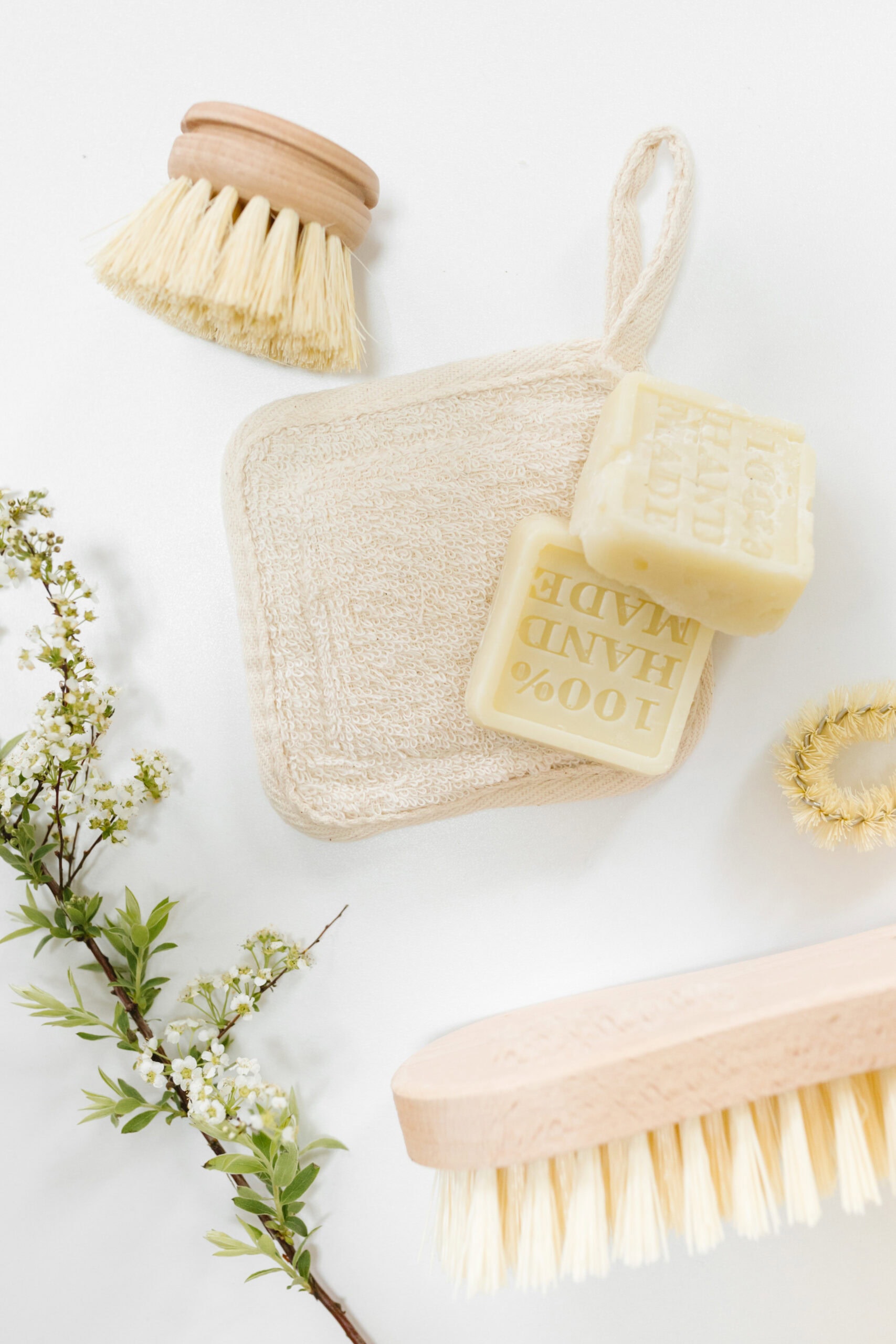 A flat lay of natural cleaning supplies, including a wooden scrub brush, a beige cleaning cloth, a bar of soap marked "100% hand made," and a sprig of delicate white flowers on a clean white background.