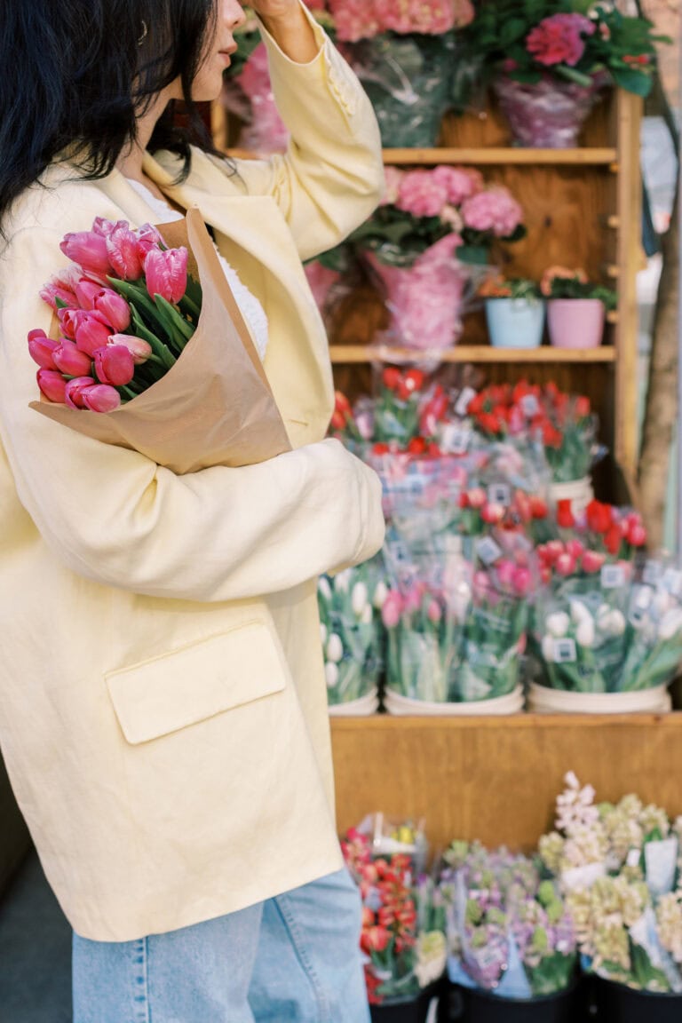 A woman in a pale yellow coat holding a bouquet of vibrant pink tulips wrapped in paper, standing in front of a flower market filled with colorful blooms in buckets and pots.