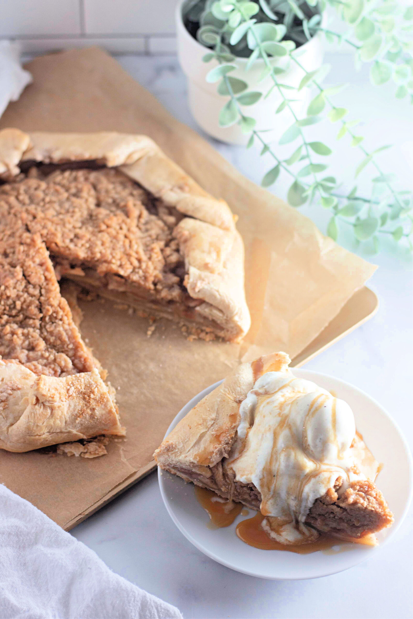 A freshly baked apple crumb galette with a golden, rustic crust and a streusel topping, sitting on a parchment-lined baking sheet. A slice has been removed and is served on a white plate with a scoop of ice cream.