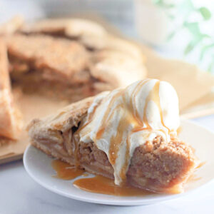 A close-up of a slice of apple crumb galette topped with vanilla ice cream and caramel drizzle. The filling is packed with tender apples, and the crumb topping is perfectly golden.
