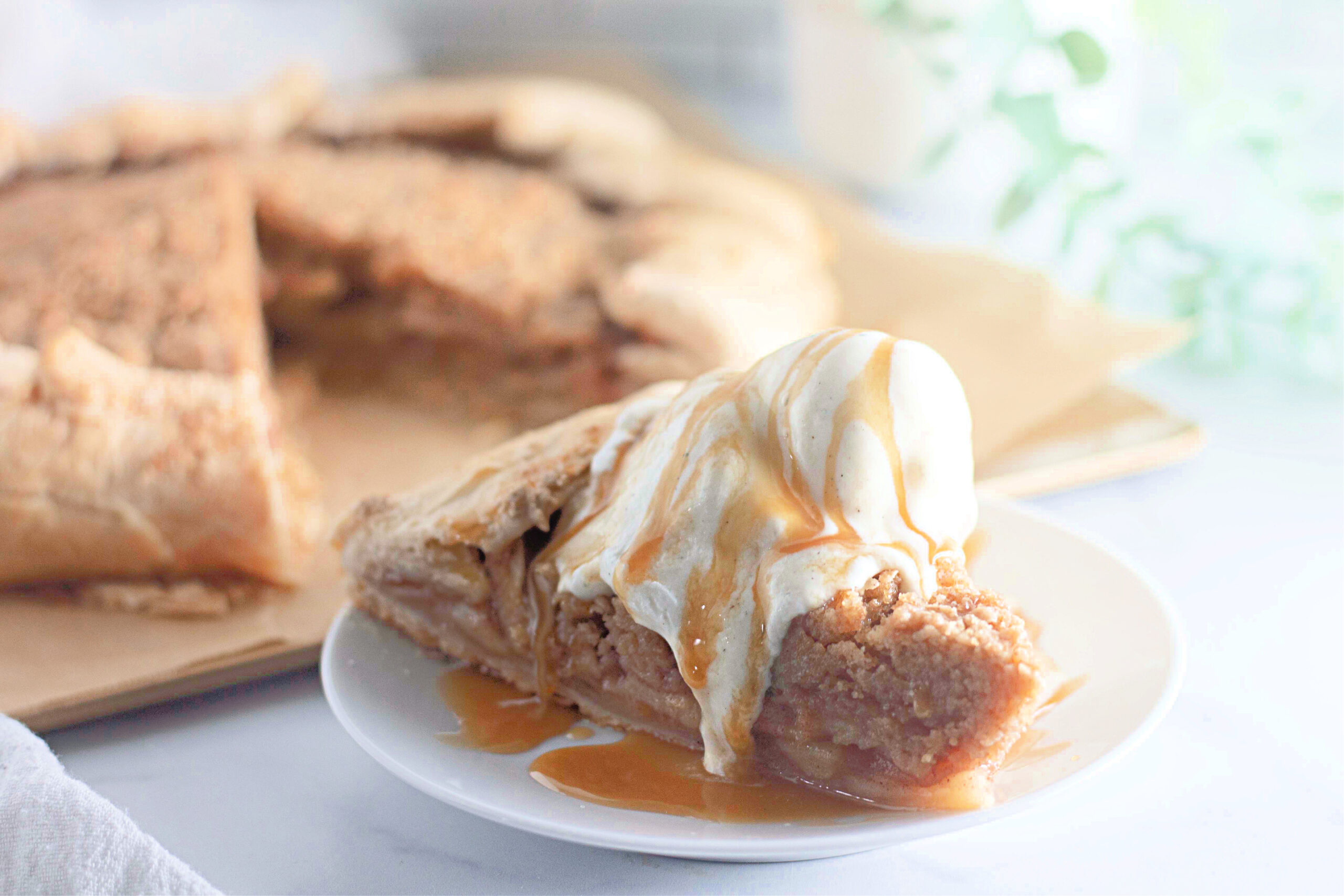 A close-up of a slice of apple crumb galette topped with vanilla ice cream and caramel drizzle. The filling is packed with tender apples, and the crumb topping is perfectly golden.