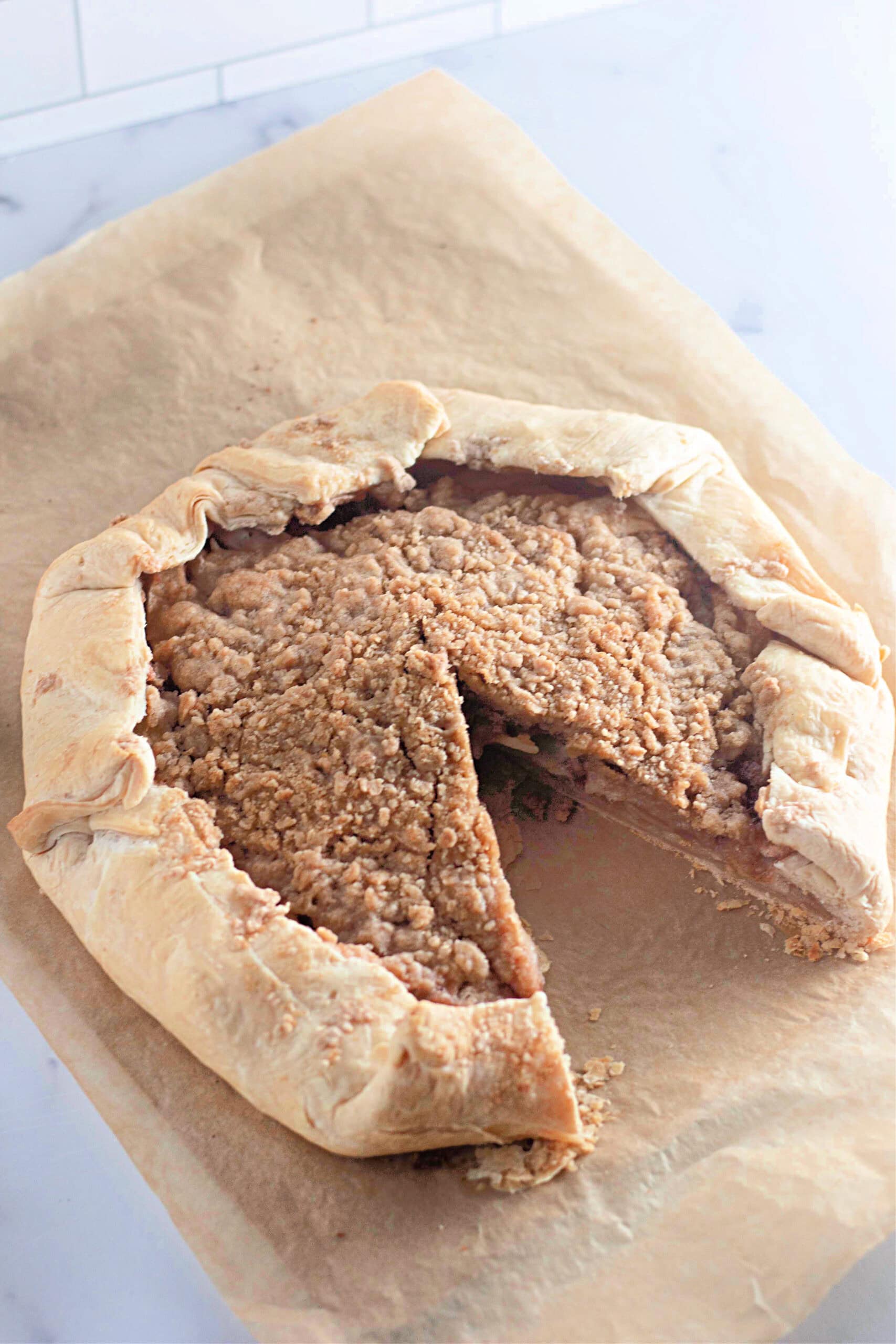 A whole apple crumb galette with a rustic, folded crust and a buttery crumb topping, baked on parchment paper. A slice has been removed, revealing the spiced apple filling inside.