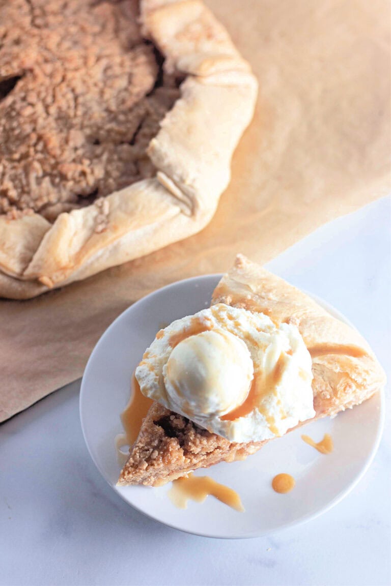 A slice of apple crumb galette on a plate, topped with a scoop of vanilla ice cream and drizzled with caramel sauce. The galette is in the background, showing its rustic, flaky crust.