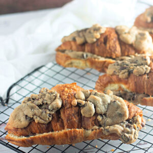 A golden-brown croissant topped with chunks of baked chocolate chip cookie dough, resting on a black wire cooling rack. The background features a soft white cloth and a pink-toned surface.