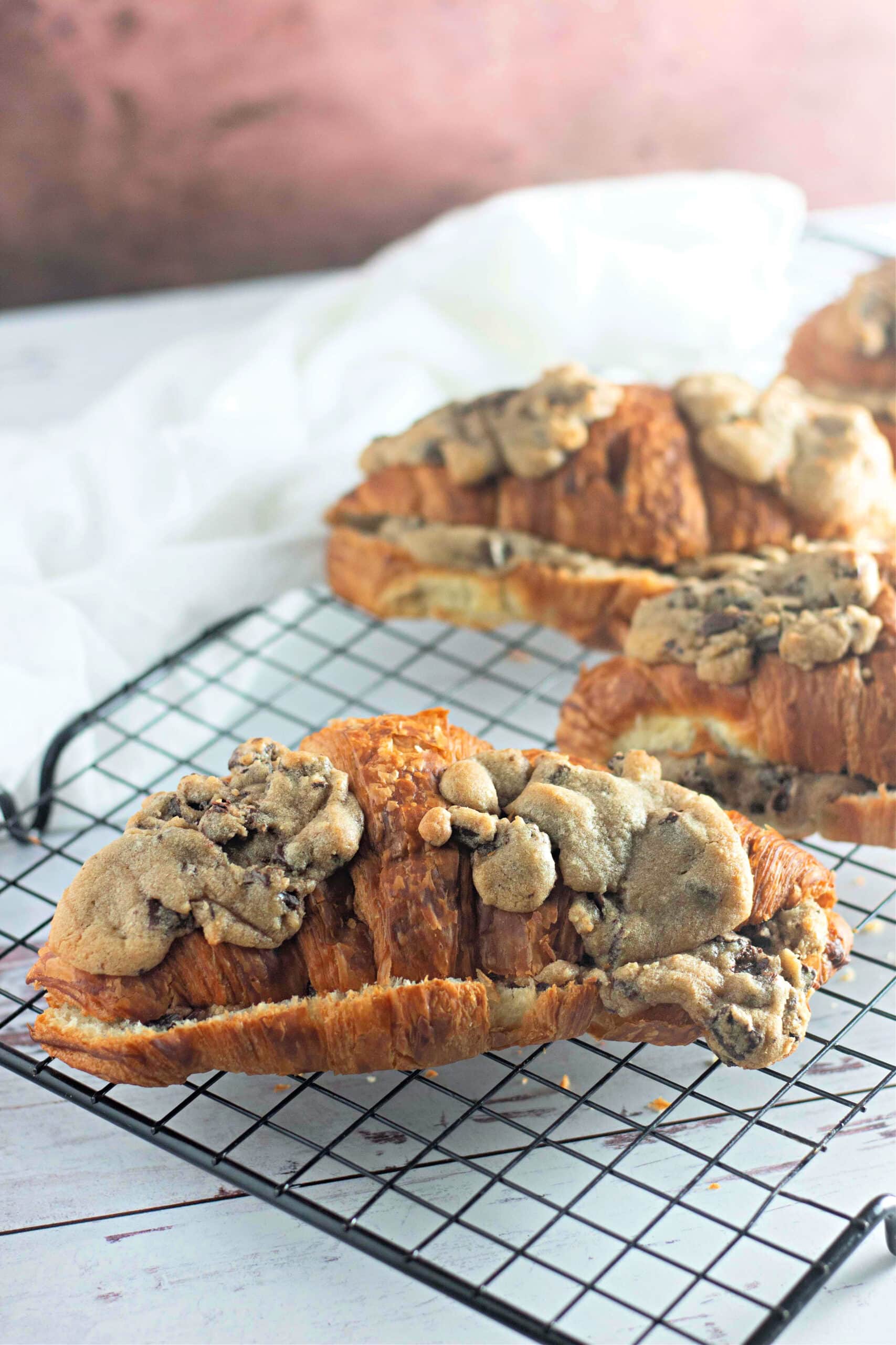 A golden-brown croissant topped with chunks of baked chocolate chip cookie dough, resting on a black wire cooling rack. The background features a soft white cloth and a pink-toned surface.