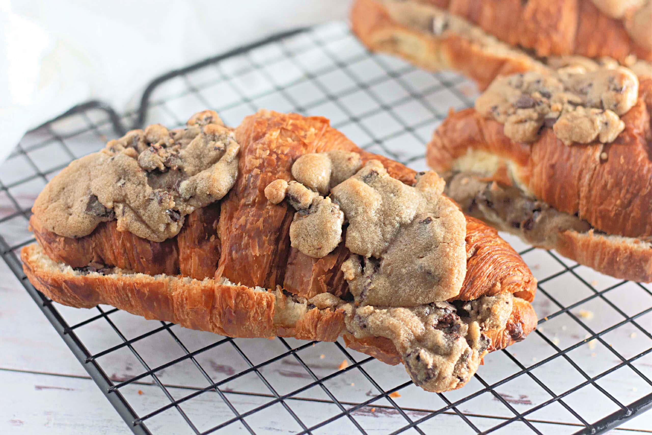A close-up of a flaky croissant with gooey chocolate chip cookie dough baked on top and inside, set on a wire rack. The melted chocolate chips are visible in the cookie topping.