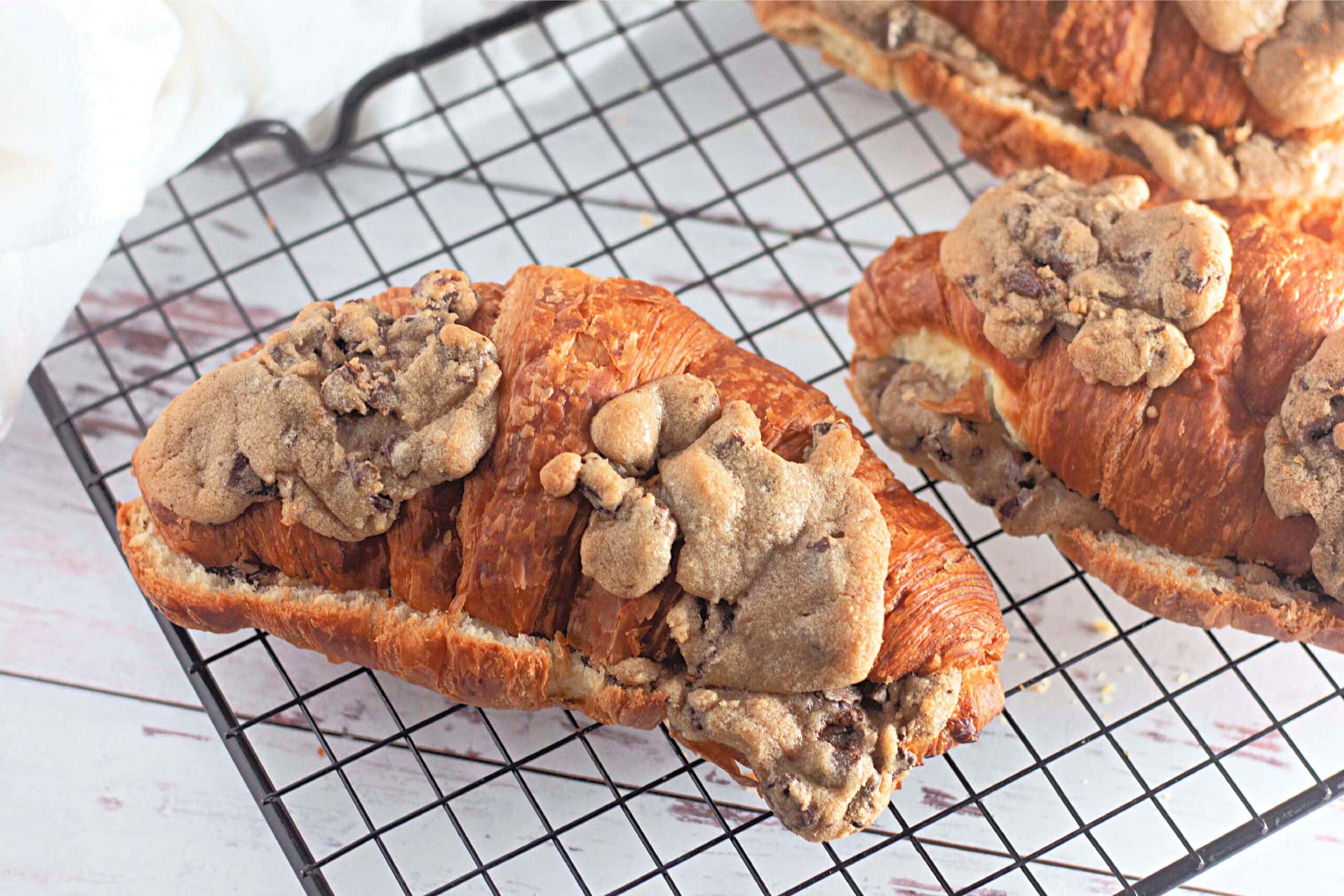 Several croissants with baked chocolate chip cookie dough on top, cooling on a black wire rack. The flaky layers of the croissants contrast with the slightly crisp cookie dough.