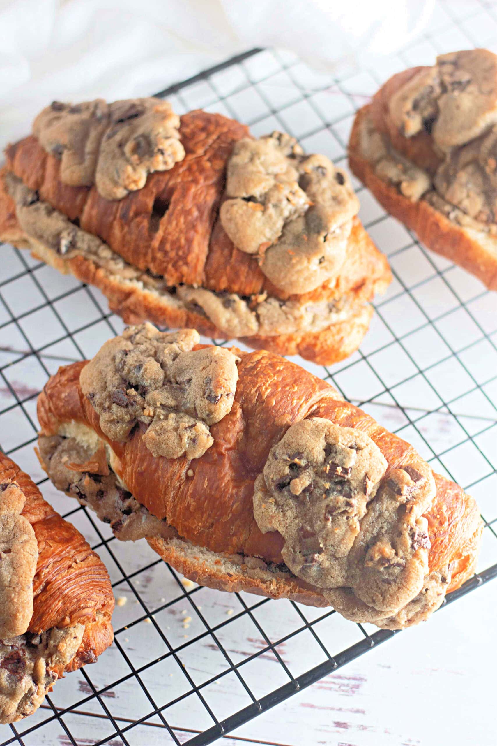 A top-down view of multiple chocolate chip cookie croissants arranged on a black wire rack. The golden, crispy croissants are generously topped with slightly melted, baked cookie dough.
