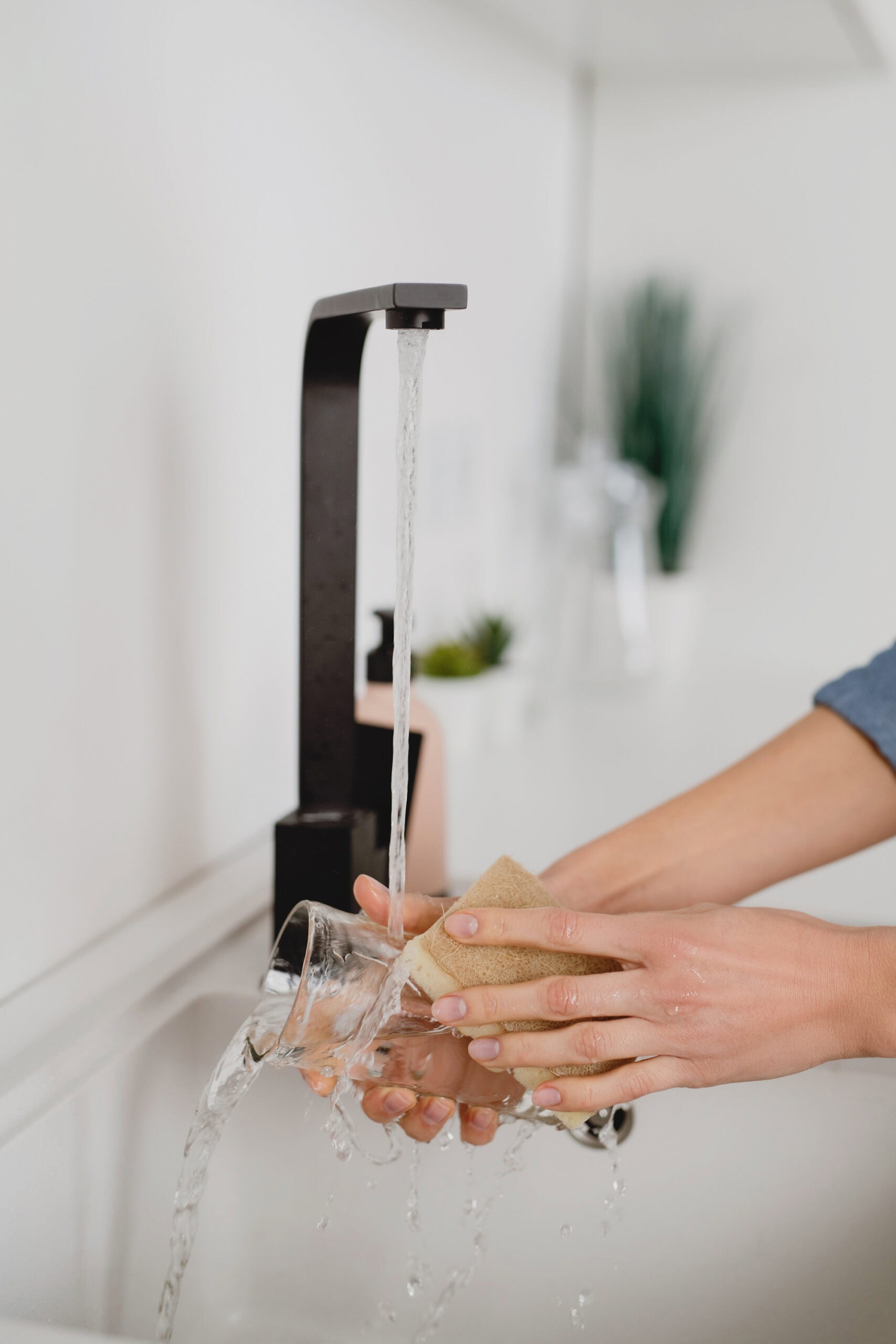 A person washes a glass cup under a running faucet, scrubbing it with a sponge as water flows over the sides.