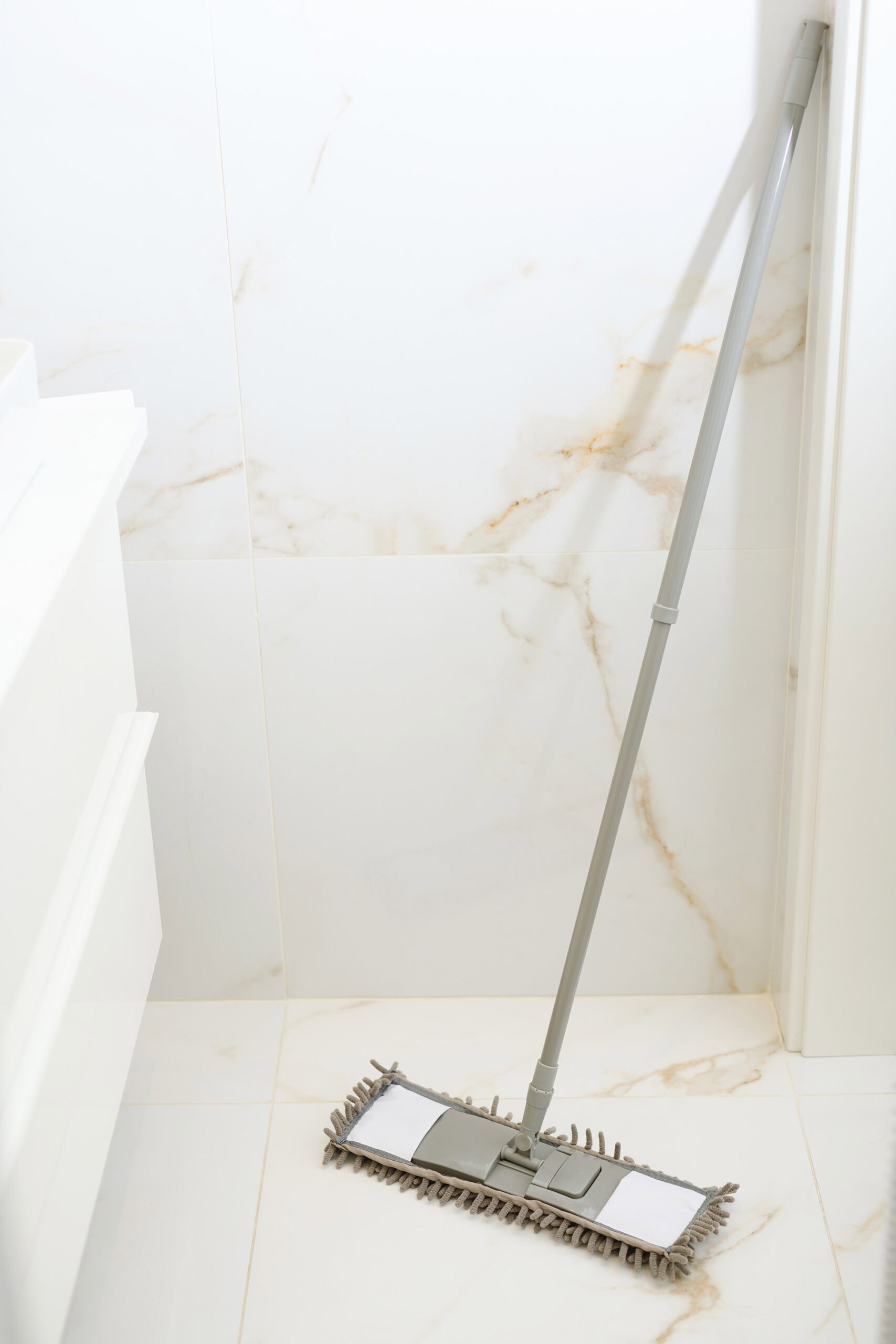 A modern, gray mop with a microfiber pad leans against a white marble wall in a bathroom, resting on a clean, polished floor.