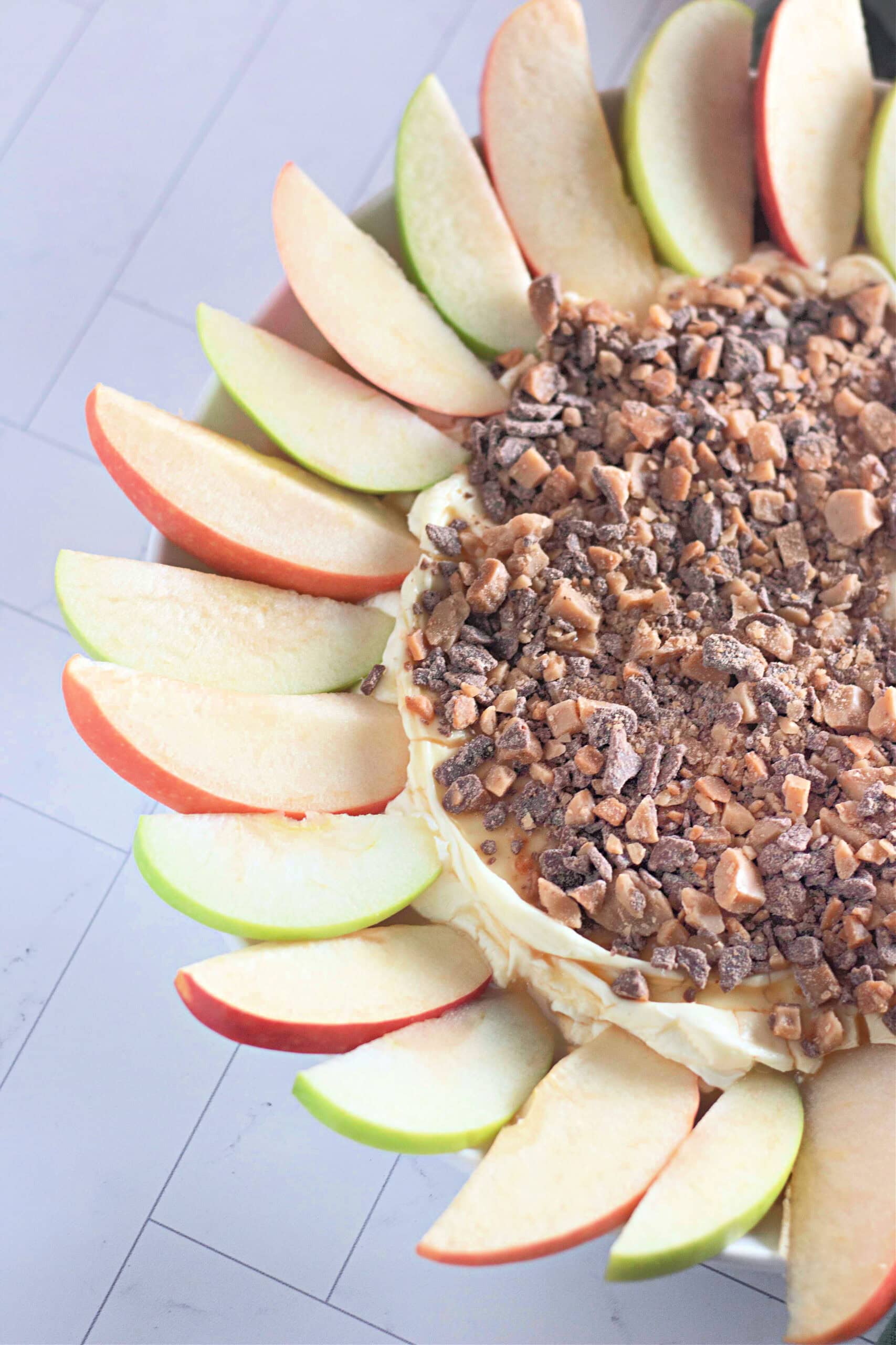 A creamy toffee apple dip topped with crushed toffee bits, surrounded by red and green apple slices arranged in a circular pattern on a white plate.