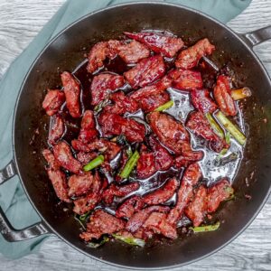 Overhead view of keto Mongolian beef cooking in a black Staub pan, featuring tender strips of beef glazed in a glossy, low-carb soy-based sauce with green onions.