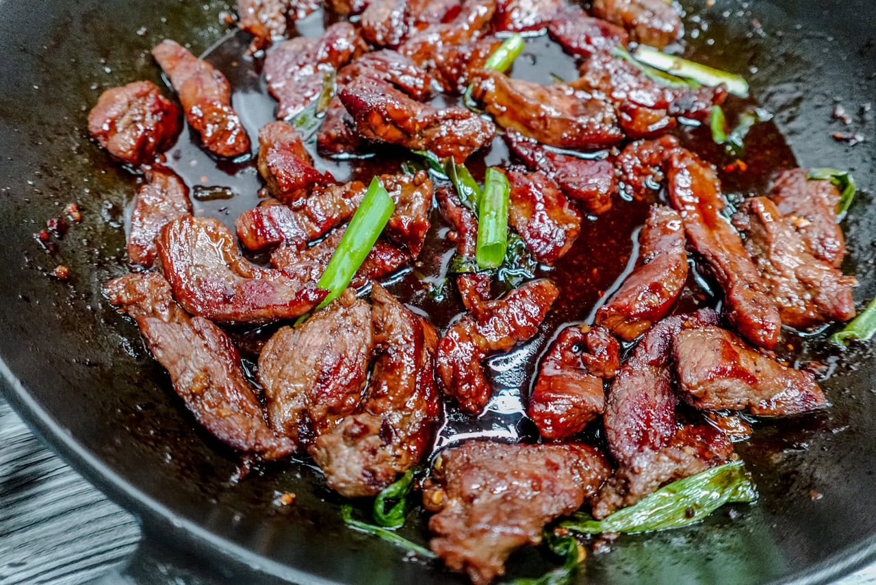 Close-up side angle of keto-friendly Mongolian beef in a cast iron skillet, highlighting the caramelized beef strips and vibrant green onions in a rich, sticky sauce.