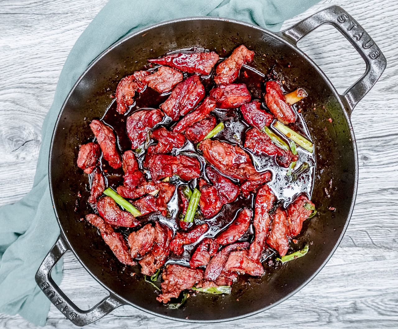 Top-down image of keto Mongolian beef served in a black Staub skillet with a rustic green napkin, showing off the deep caramel color and fresh green onion slices.