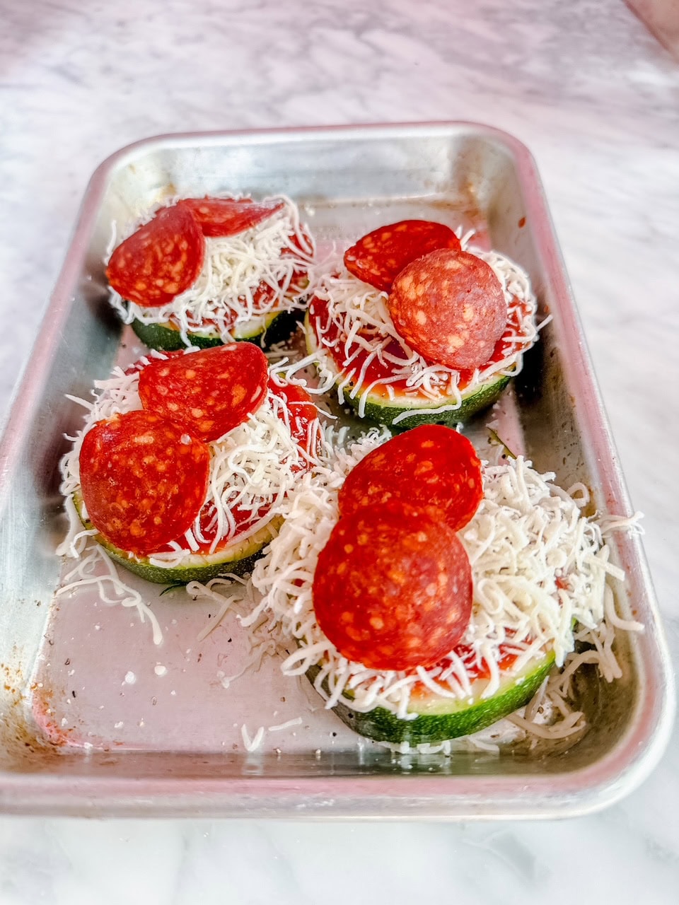 Unbaked zucchini pepperoni pizzas on a silver baking tray, topped with marinara sauce, shredded mozzarella cheese, and mini pepperoni slices, ready for the oven.