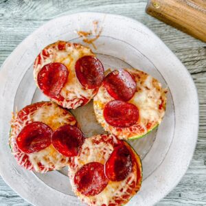 Overhead view of four zucchini pizza rounds on a white plate, each topped with melted cheese and golden pepperoni slices.