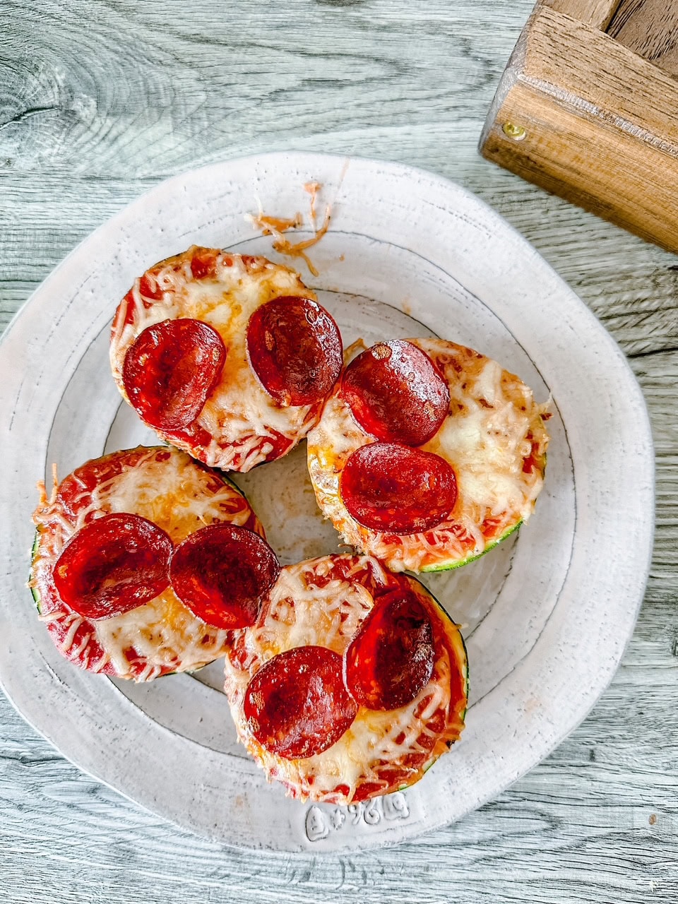 Overhead view of four zucchini pizza rounds on a white plate, each topped with melted cheese and golden pepperoni slices.