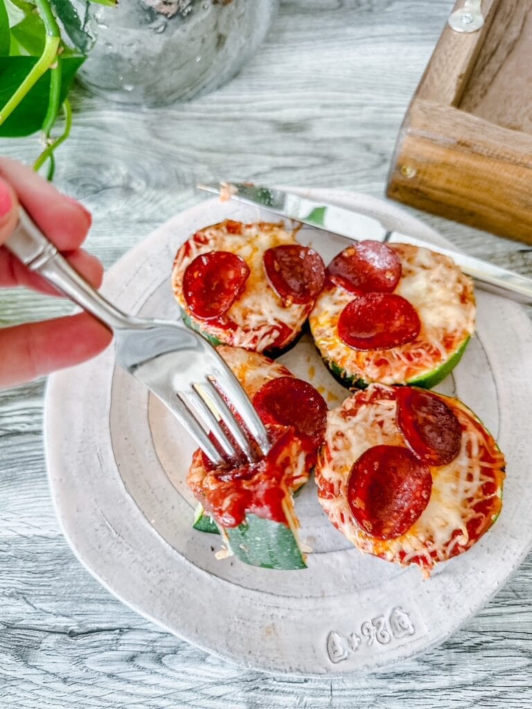 A fork lifting a bite of zucchini pepperoni pizza off a plate, showing the layers of zucchini, tomato sauce, melty cheese, and pepperoni—perfect for a low-carb snack or appetizer.