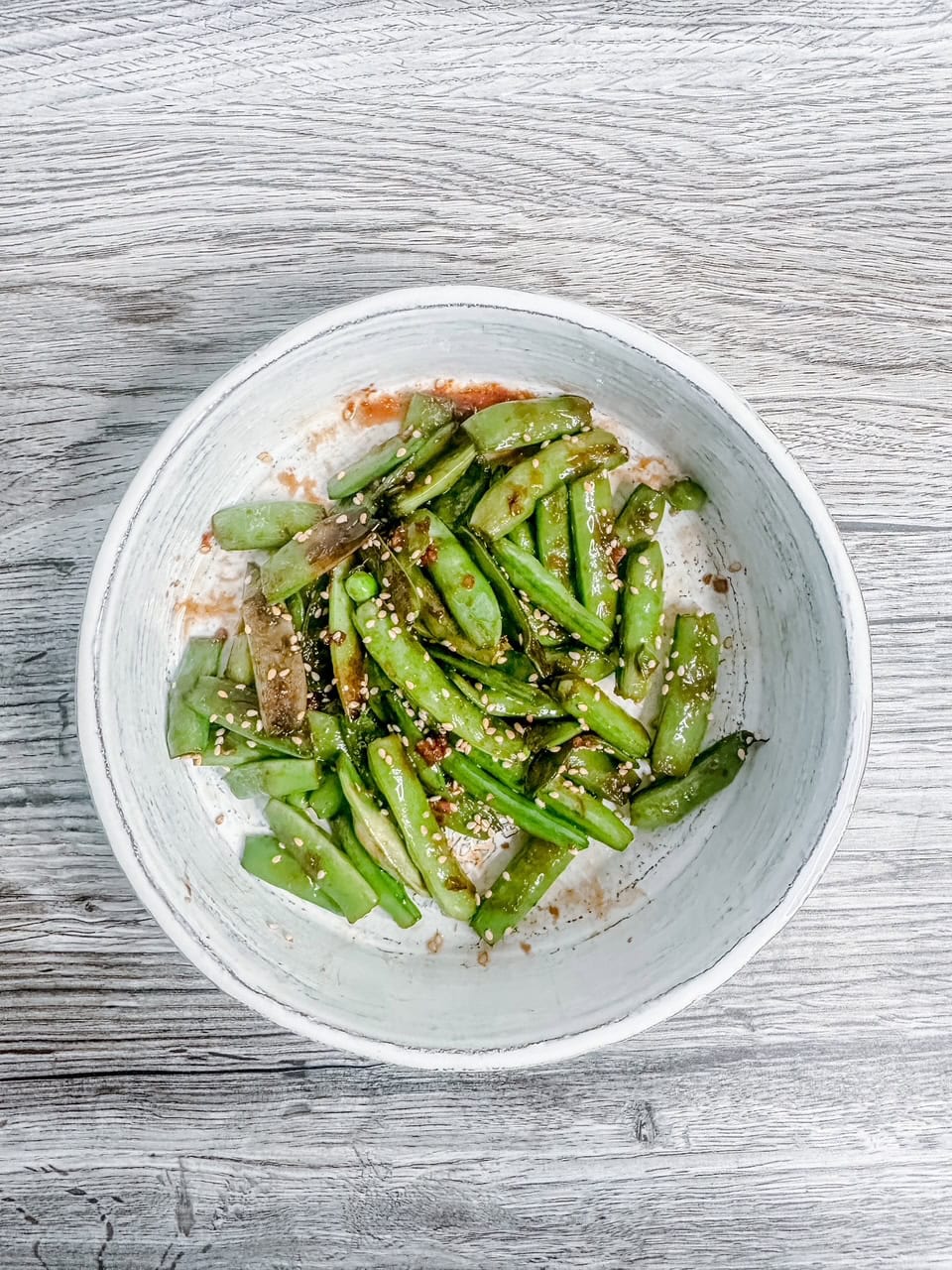 Wide overhead shot of sesame snow peas in a rustic white bowl on a grey wooden surface, capturing a clean, minimalist food presentation.