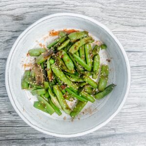A white ceramic bowl filled with vibrant green sautéed snow peas, garnished with sesame seeds and bits of caramelized garlic, set on a light woodgrain background.