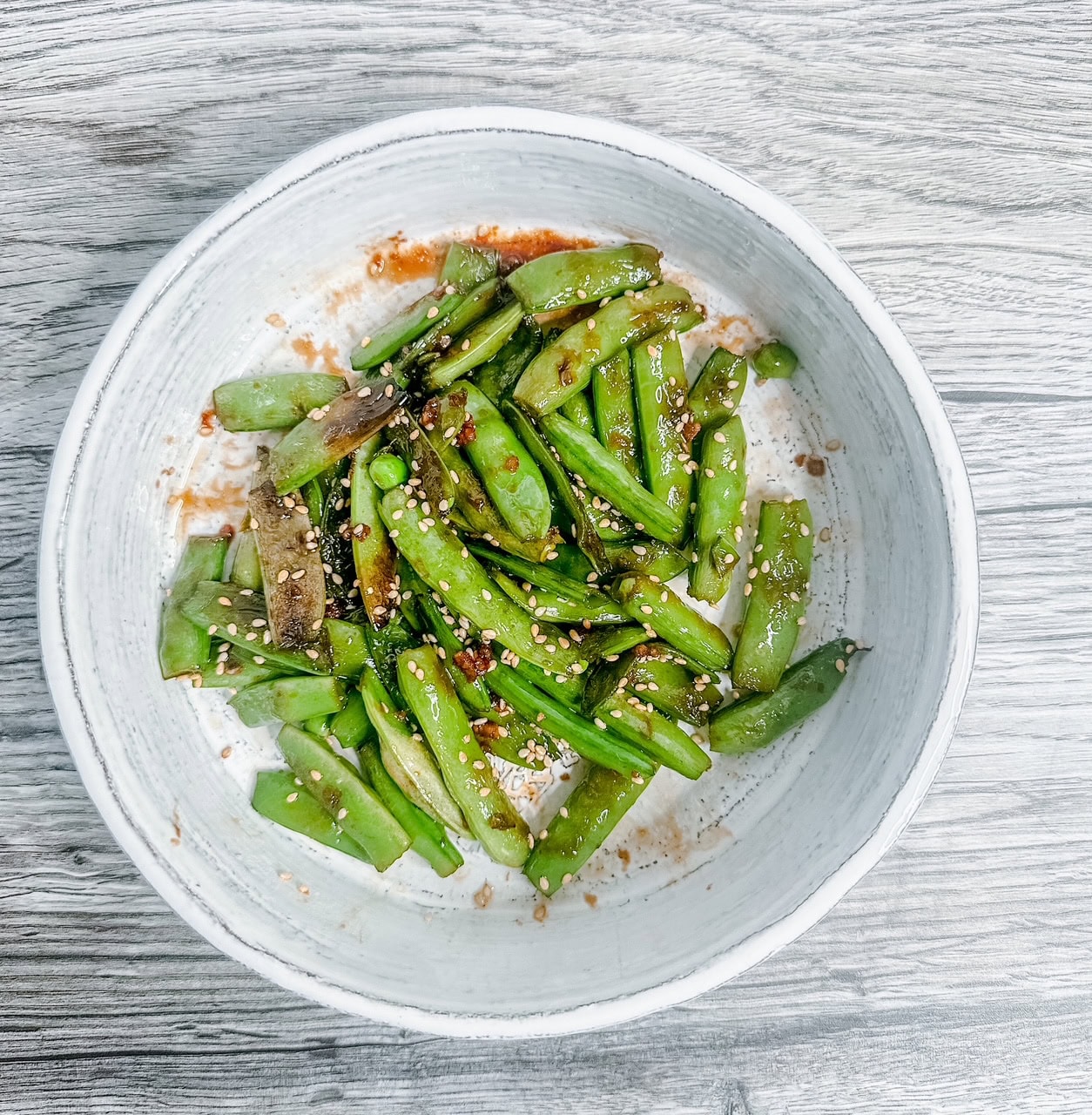 A white ceramic bowl filled with vibrant green sautéed snow peas, garnished with sesame seeds and bits of caramelized garlic, set on a light woodgrain background.
