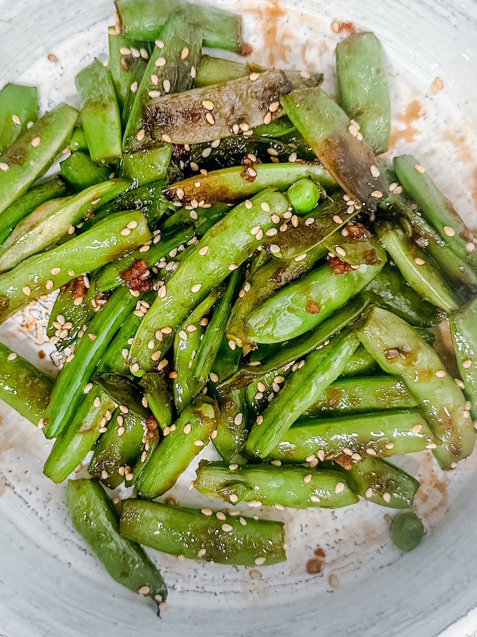 Close-up view of sesame snow peas coated in a glossy sauce, garnished with toasted sesame seeds, highlighting their texture and vibrant green color.