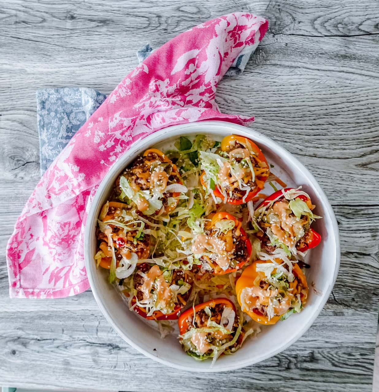 Top-down view of a white dish filled with colorful mini bell peppers stuffed with seasoned ground beef, shredded lettuce, sesame seeds, melted cheese, and drizzled with Big Mac-style sauce. A pink and white floral napkin is tucked beside the bowl on a rustic wood surface.
