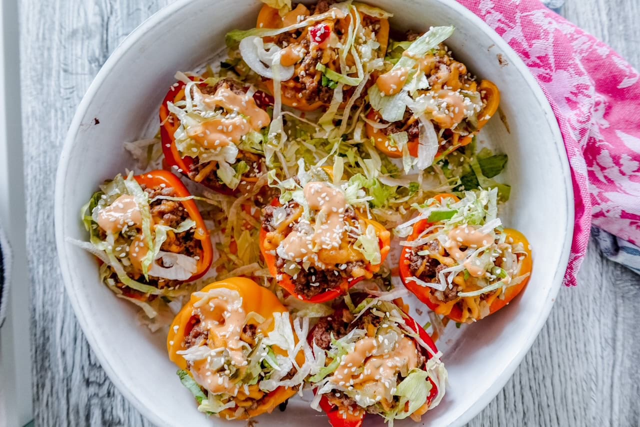 Close-up top view of Big Mac stuffed peppers in a round white dish, showcasing the vibrant red, orange, and yellow pepper halves topped with shredded lettuce, cheese, sesame seeds, and creamy sauce.