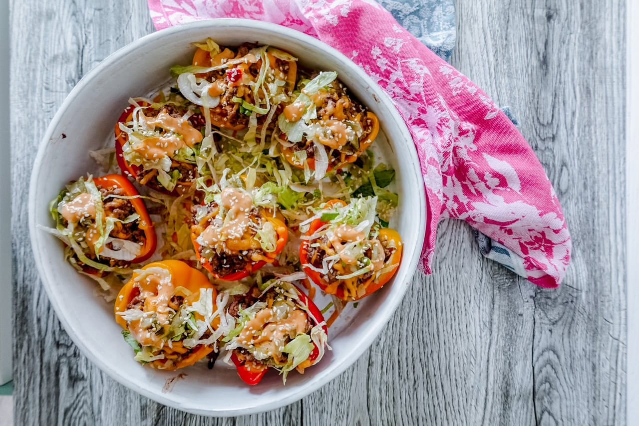Bright overhead photo of stuffed mini peppers arranged in a white bowl, garnished generously with shredded lettuce and Big Mac sauce, with a pink floral cloth partially surrounding the dish.