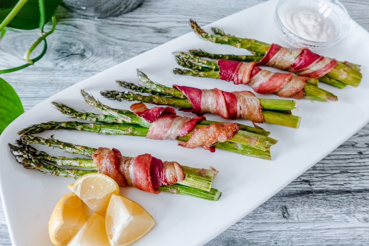 Bacon-wrapped asparagus bundles arranged on a white rectangular platter with lemon wedges on the side and a small bowl of creamy dipping sauce.