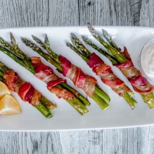 Bacon-wrapped asparagus bundles arranged on a white rectangular platter with lemon wedges on the side and a small bowl of creamy dipping sauce.