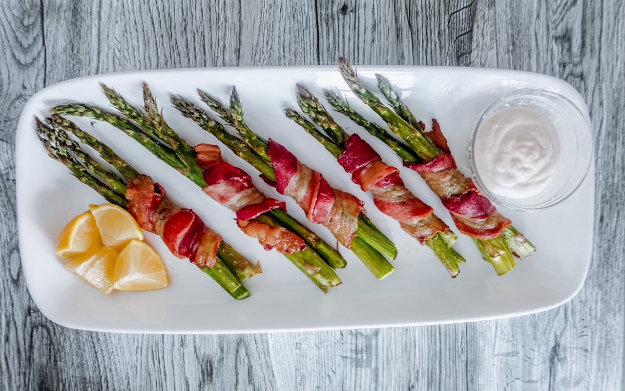 Bacon-wrapped asparagus bundles arranged on a white rectangular platter with lemon wedges on the side and a small bowl of creamy dipping sauce.