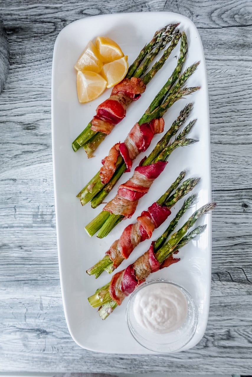 Top-down view of five bacon-wrapped asparagus bundles on a white serving platter with lemon wedges and a bowl of dip.