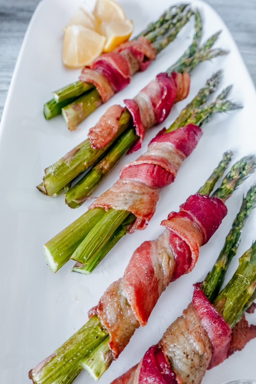 Angled overhead view of bacon-wrapped asparagus bundles garnished with lemon wedges, showcasing the crisp tips of the asparagus.