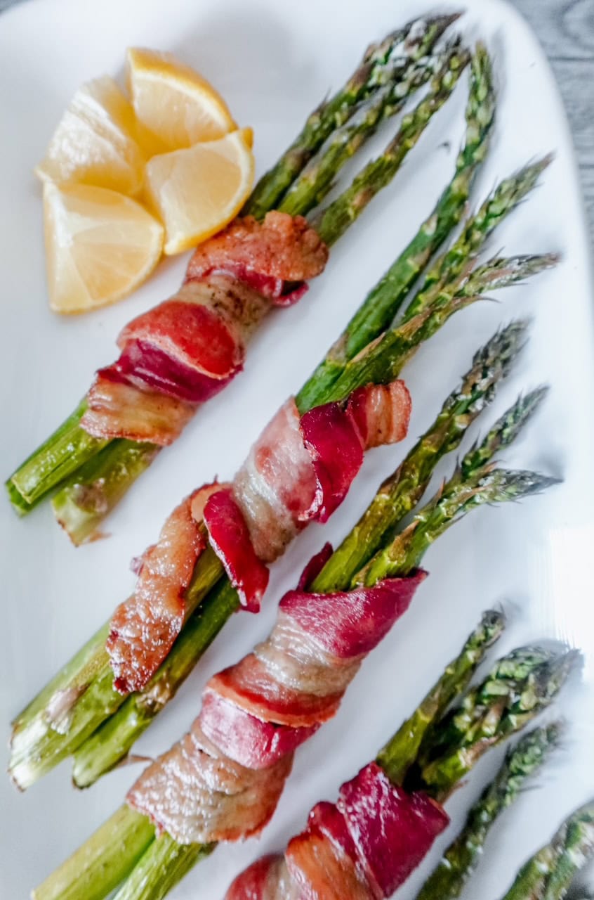 Close-up of crispy bacon-wrapped asparagus bundles with visible texture and golden edges, served with lemon slices on a white platter.