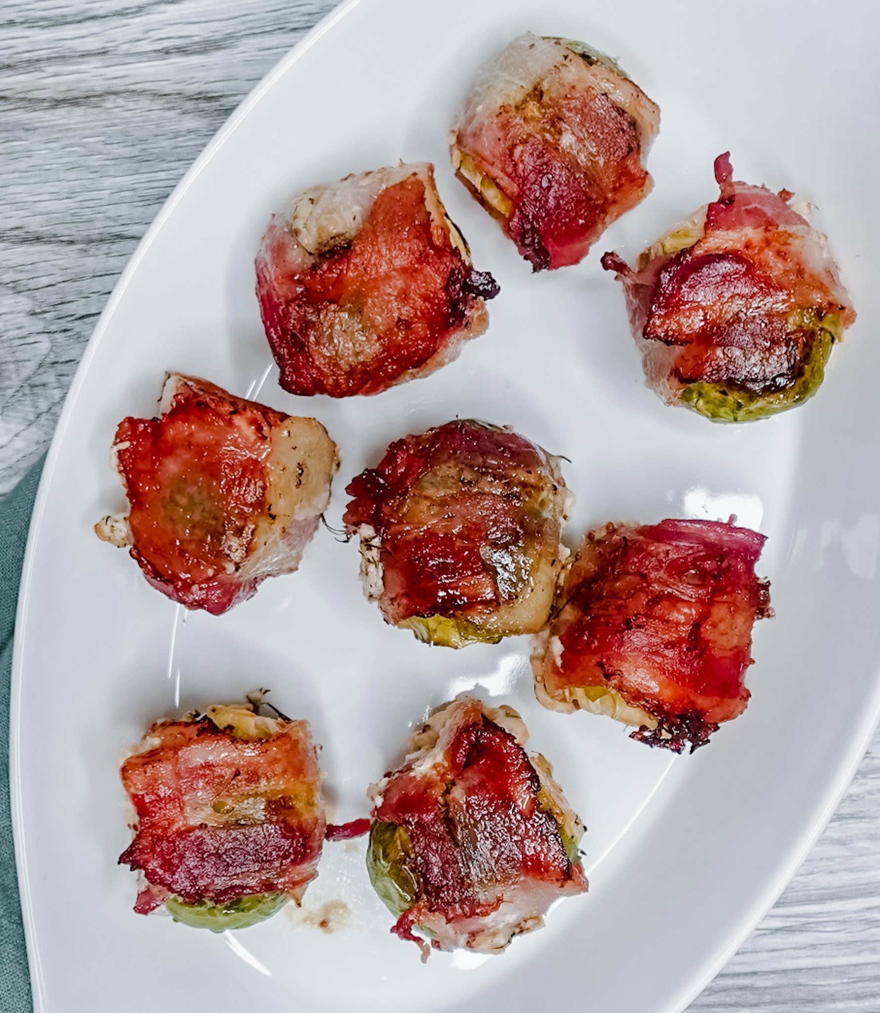 Overhead close-up of bacon-wrapped Brussels sprouts, evenly spaced on a white platter with charred and golden crisped edges.