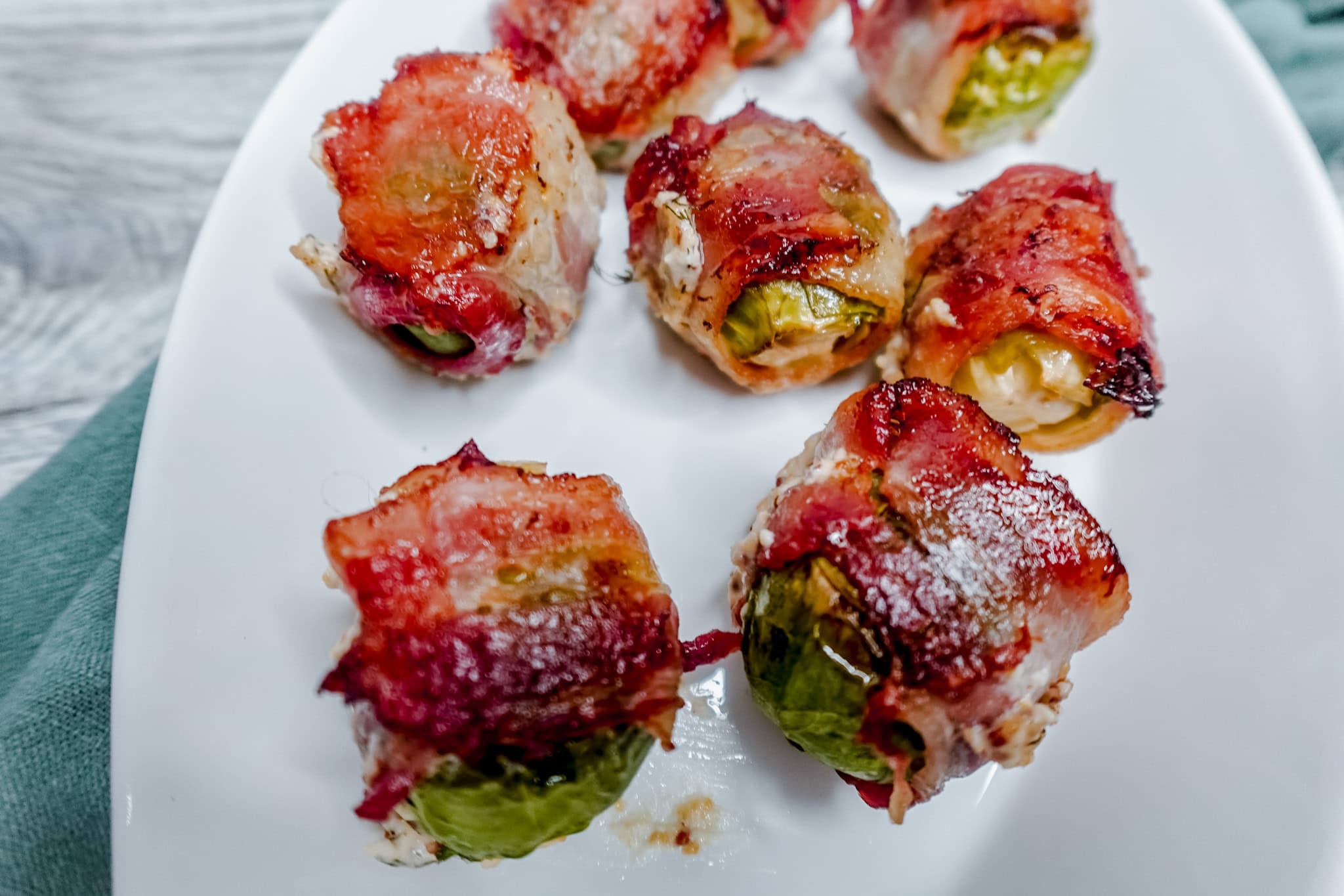Bacon-wrapped Brussels sprouts on a white plate, showing detail of the melted cheese and browned bacon wrapping each sprout.