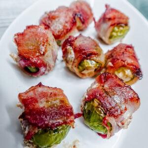 Close-up of crispy bacon-wrapped Brussels sprouts on a white platter, showcasing the golden brown bacon and tender roasted sprouts.