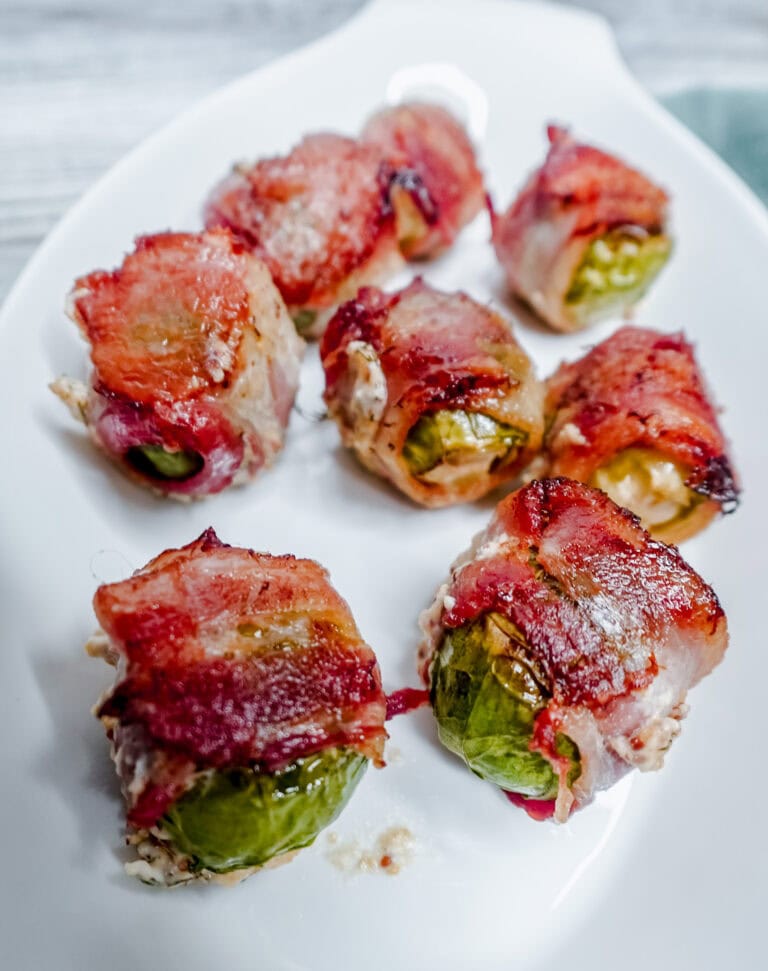 Close-up of crispy bacon-wrapped Brussels sprouts on a white platter, showcasing the golden brown bacon and tender roasted sprouts.