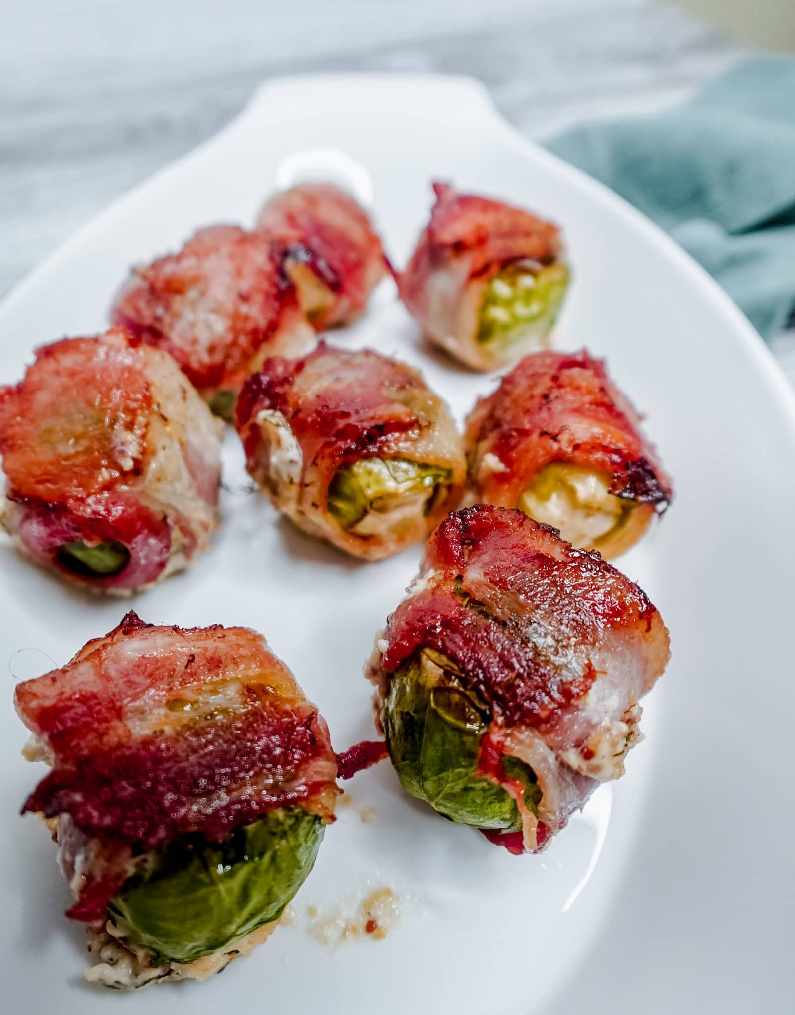 Top-down view of eight bacon-wrapped Brussels sprouts arranged in a curved white dish, with a green cloth napkin to the side.