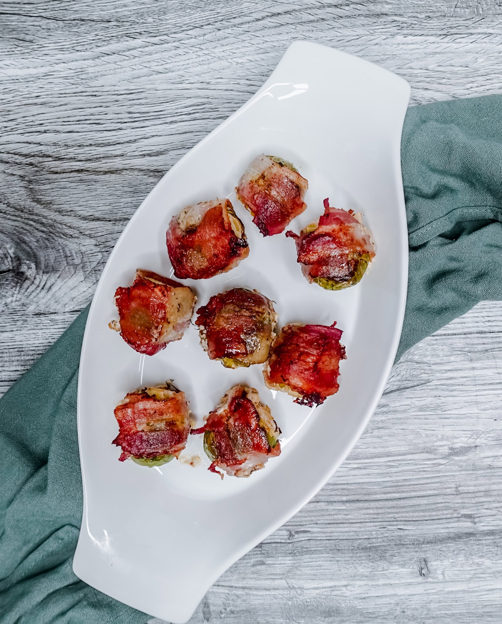 Top-down view of eight bacon-wrapped Brussels sprouts arranged in a curved white dish, with a green cloth napkin to the side.