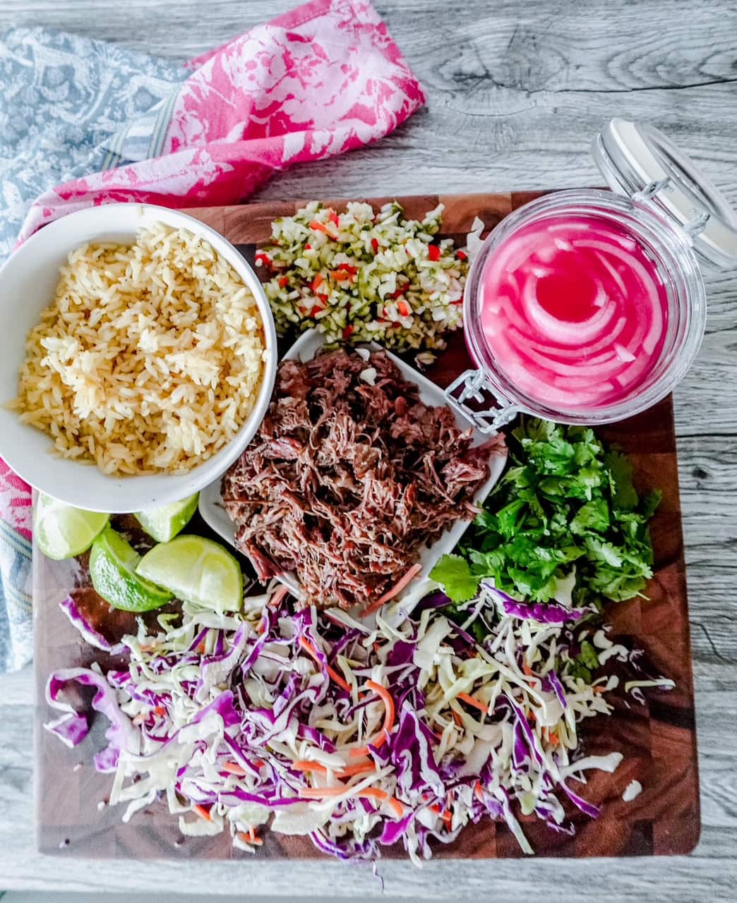 Flat lay of a vibrant taco board featuring seasoned rice, shredded short rib, pickled onions, lime wedges, chopped herbs, slaw, and colorful diced veggies arranged on a wood board.