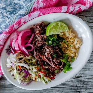 Finished short rib taco bowl with rice, pickled onions, shredded beef, cilantro, and slaw served in a white bowl with a wedge of lime on the side.