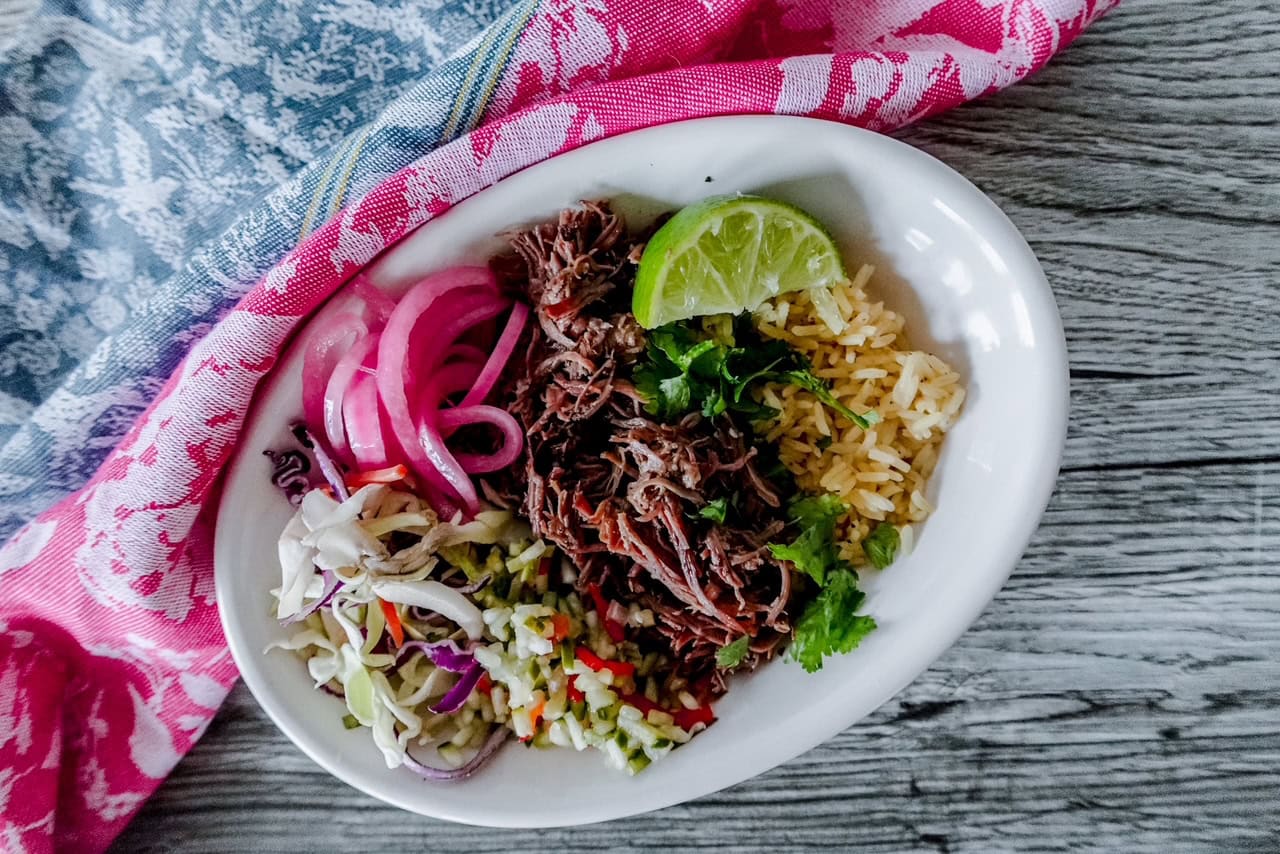 Finished short rib taco bowl with rice, pickled onions, shredded beef, cilantro, and slaw served in a white bowl with a wedge of lime on the side.